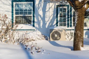 Dual fuel heat pump outdoor unit running in winter beside a snow-covered home