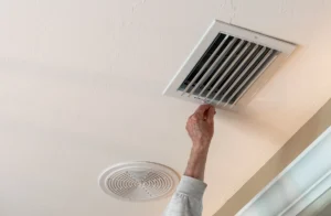 Ceiling return air grille with a homeowner lifting the grille cover during an HVAC airflow and filter check.